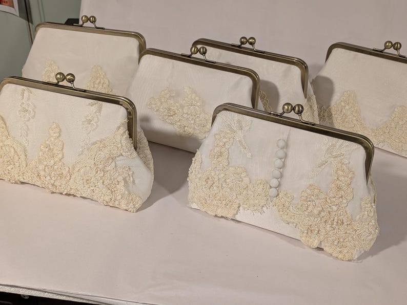May include: Several off-white clutch purses with metal clasps are displayed. The purses are adorned with delicate, cream-colored lace detailing. Some purses have a row of small, white buttons. The purses are arranged on a white surface.