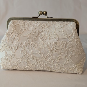 May include: Ivory lace clutch with a bronze clasp. The clutch features an intricate floral lace pattern over a satin-like fabric. The clasp is a classic kiss-lock style with two small, round knobs.