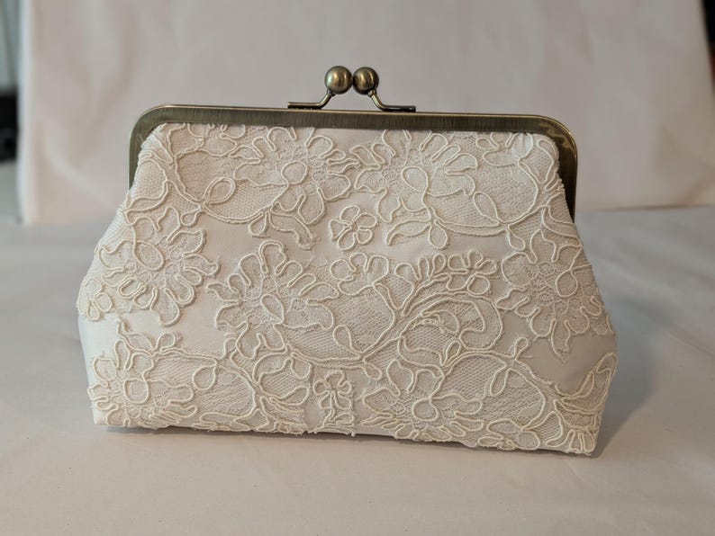 May include: Ivory lace clutch with a bronze clasp. The clutch features an intricate floral lace pattern over a satin-like fabric. The clasp is a classic kiss-lock style with two small, round knobs.