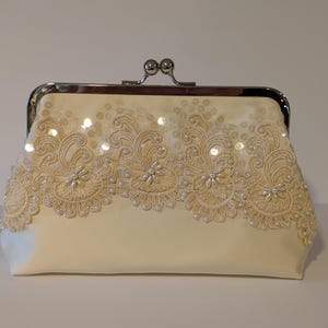 May include: Ivory-colored clutch with a silver clasp. The front of the bag is adorned with intricate lace and sequin detailing. The lace design features floral patterns and is embellished with small pearls.