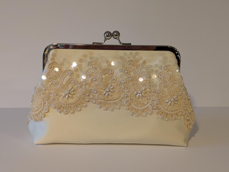 May include: Ivory-colored clutch with a silver clasp. The front of the bag is adorned with intricate lace and sequin detailing. The lace design features floral patterns and is embellished with small pearls.