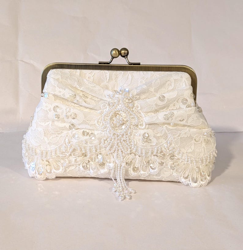 May include: Ivory lace clutch with a brass clasp. The clutch features intricate lace detailing, embellished with beads and sequins. A beaded tassel hangs from the center, adding a touch of elegance. The frame is a warm brass color.