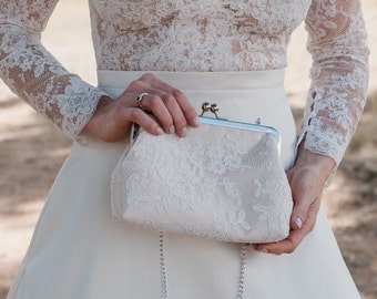 May include: A cream-colored clutch bag featuring an intricate floral lace overlay, a silver-toned metal frame, and a kiss-lock clasp closure. The accessory includes a thin silver-toned metal chain strap and has a structured, rectangular shape.