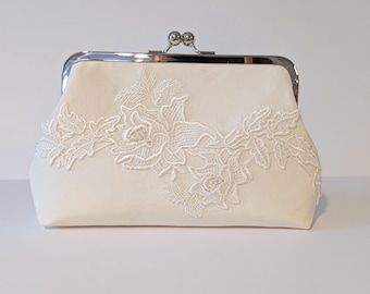 May include: Ivory clutch with a silver clasp and intricate white floral lace detailing. The rectangular bag features a kiss-lock closure and a delicate, romantic design. A perfect accessory for special occasions.