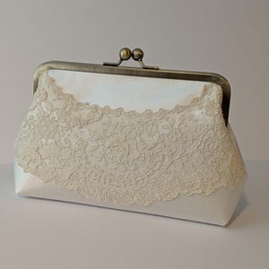 May include: A white satin clutch with a decorative ivory lace overlay. The clutch features a vintage-style metal clasp and frame. The lace design adds a delicate, feminine touch to the accessory.