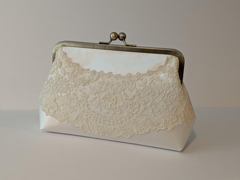 May include: A white satin clutch with a decorative ivory lace overlay. The clutch features a vintage-style metal clasp and frame. The lace design adds a delicate, feminine touch to the accessory.
