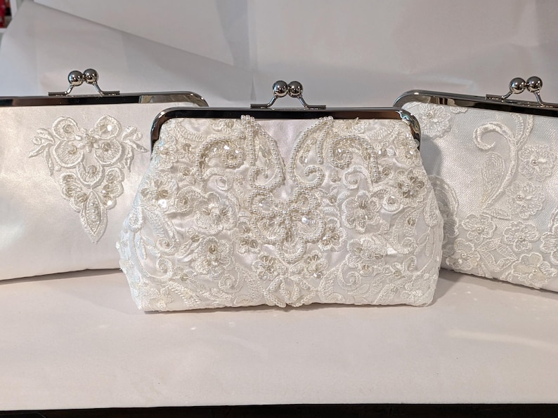 May include: Three white bridal clutch purses with silver-toned metal clasps. The purses are embellished with intricate white lace and beadwork. The central purse features a detailed floral design, while the others have unique lace patterns.