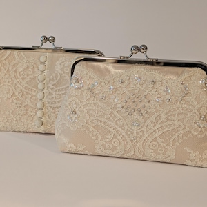 May include: Two ivory lace clutch purses with silver-toned metal clasps. One purse has a row of buttons down the front. The other is embellished with small, sparkling beads. The purses are on a white surface.