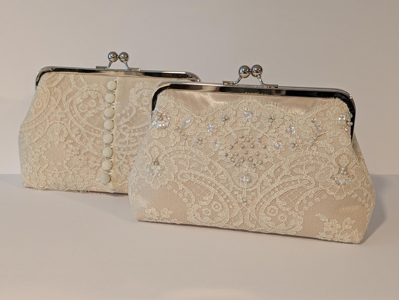 May include: Two ivory lace clutch purses with silver-toned metal clasps. One purse has a row of buttons down the front. The other is embellished with small, sparkling beads. The purses are on a white surface.