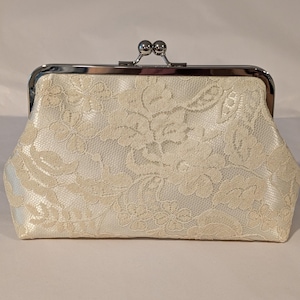 May include: Ivory lace clutch with a silver clasp. The clutch features a floral lace pattern over a satin fabric. The clasp is a silver metal frame with a kiss-lock closure.