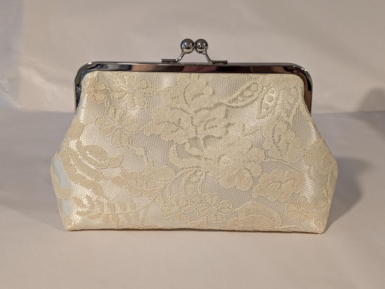 May include: Ivory lace clutch with a silver clasp. The clutch features a floral lace pattern over a satin fabric. The clasp is a silver metal frame with a kiss-lock closure.