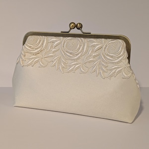 May include: Ivory clutch with a decorative floral lace trim. The clasp is a vintage-style bronze color. The clutch is a soft, rectangular shape, perfect for carrying essentials. The lace trim features a repeating pattern of roses and leaves.