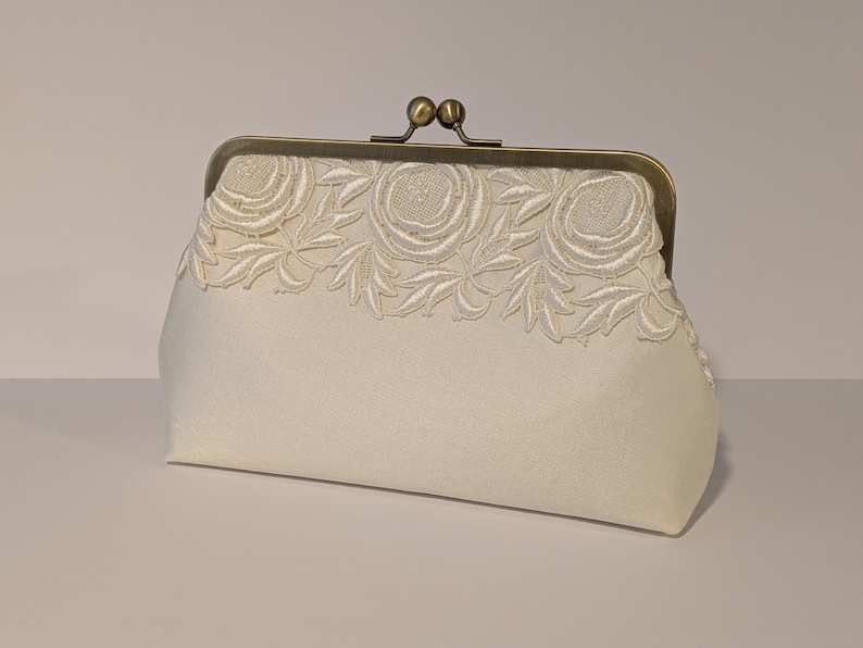 May include: Ivory clutch with a decorative floral lace trim. The clasp is a vintage-style bronze color. The clutch is a soft, rectangular shape, perfect for carrying essentials. The lace trim features a repeating pattern of roses and leaves.