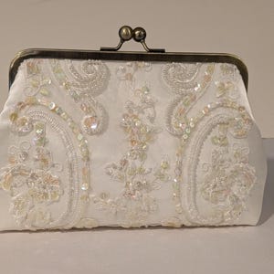 May include: Ivory satin clutch with ornate floral embroidery and iridescent sequins. The clasp is a dark bronze color. The bag is a rectangular shape with a kiss-lock closure. A formal accessory.