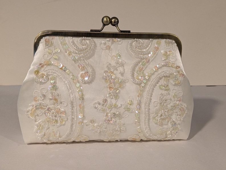 May include: Ivory satin clutch with ornate floral embroidery and iridescent sequins. The clasp is a dark bronze color. The bag is a rectangular shape with a kiss-lock closure. A formal accessory.