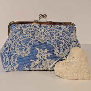 May include: A blue clutch with a silver clasp and white lace overlay. A cream-colored heart-shaped decoration with lace detailing sits beside the clutch. The clutch and heart are likely for a wedding or special occasion.