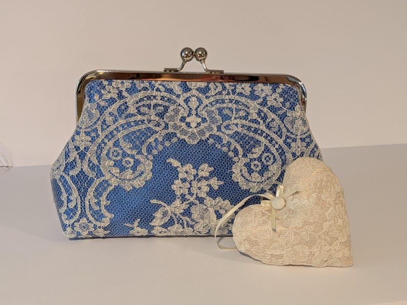 May include: A blue clutch with a silver clasp and white lace overlay. A cream-colored heart-shaped decoration with lace detailing sits beside the clutch. The clutch and heart are likely for a wedding or special occasion.