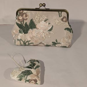 May include: A beige clutch with a floral embroidered design, featuring pink, green, and brown accents. A matching heart-shaped ornament with similar embroidery is also present. The clutch has a metal clasp.