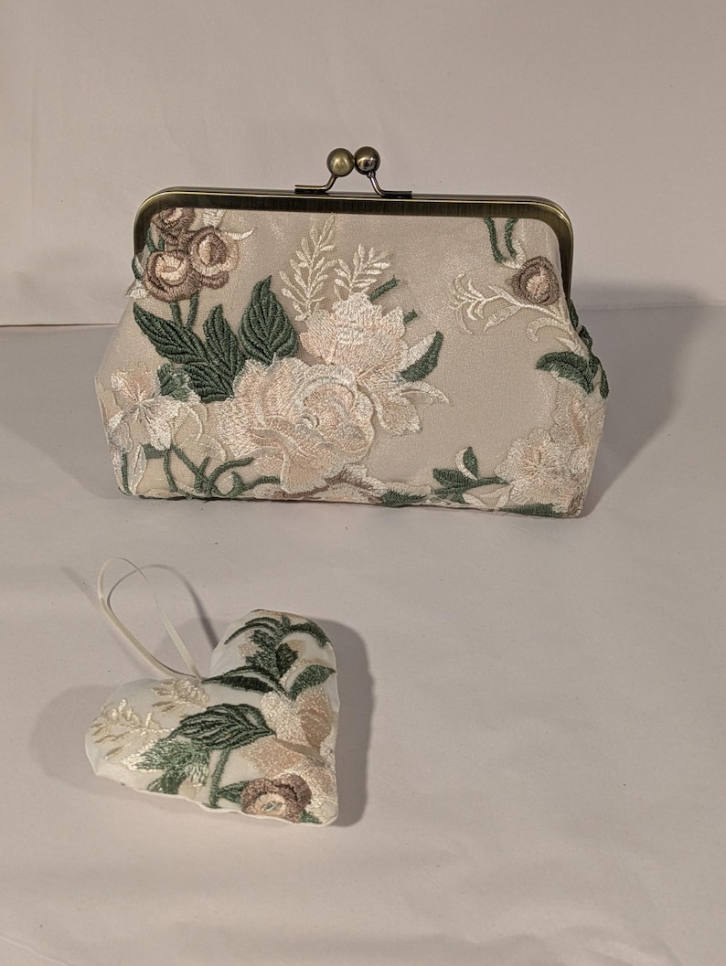 May include: A beige clutch with a floral embroidered design, featuring pink, green, and brown accents. A matching heart-shaped ornament with similar embroidery is also present. The clutch has a metal clasp.