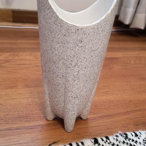 May include: A tall, speckled gray vase with a white interior. The vase has a unique, flared top and three small feet. The speckled pattern gives it a textured appearance, and it stands approximately 12 inches tall.