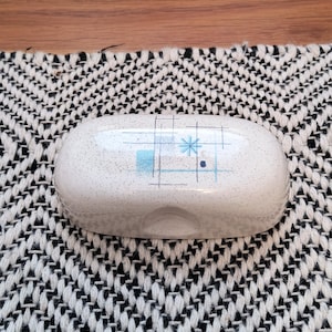 May include: White ceramic soap dish with a speckled finish and a mid-century modern design. The dish features a blue geometric pattern with a star and grid design. The dish is oval-shaped and sits on a black and white patterned surface.