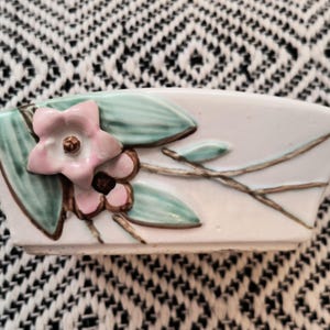 May include: A white ceramic planter with a floral design. The planter features a pink flower with a brown center, green leaves, and brown stems. The planter is set against a black and white patterned background.