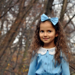 Puede incluir: Una ni&ntilde;a joven con cabello largo y casta&ntilde;o lleva un vestido azul claro con un cuello de encaje blanco y un lazo azul claro en el cabello. Est&aacute; de pie frente a un fondo borroso de &aacute;rboles.