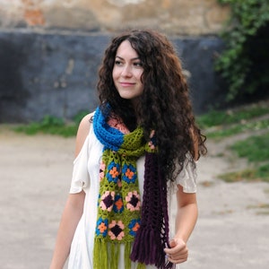 Könnte beinhalten: Eine Frau mit langen, lockigen braunen Haaren trägt ein weißes Oberteil und eine schwarze Hose. Sie trägt einen bunten Häkel-Schal mit grünen, blauen, orangefarbenen und violetten Akzenten.