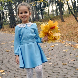 Puede incluir: Una ni&ntilde;a peque&ntilde;a con un vestido azul claro con un cuello de encaje blanco sostiene un ramo de hojas de oto&ntilde;o amarillas. Lleva medias blancas y zapatos blancos. La ni&ntilde;a est&aacute; de pie en un camino de adoquines.