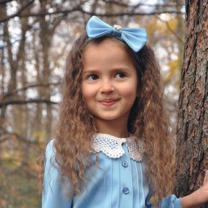 Puede incluir: Una ni&ntilde;a joven con el pelo largo y rizado casta&ntilde;o lleva un vestido azul claro con un cuello de ganchillo blanco y una diadema con lazo azul claro. Est&aacute; de pie en un bosque con &aacute;rboles y hojas en el fondo.