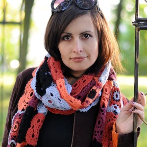 May include: A woman wearing a brown jacket with a colorful crocheted scarf. She is wearing sunglasses and has brown hair.