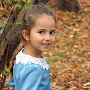 Puede incluir: Una ni&ntilde;a joven con cabello largo y casta&ntilde;o que lleva un vestido azul claro con un cuello de encaje blanco. Est&aacute; de pie en un bosque con hojas ca&iacute;das en el suelo.