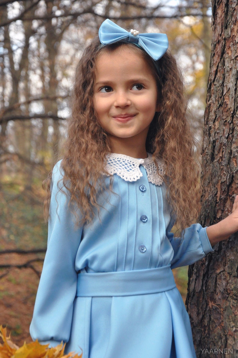 Puede incluir: Una ni&ntilde;a joven con el pelo largo y rizado casta&ntilde;o lleva un vestido azul claro con un cuello de ganchillo blanco y una diadema con lazo azul claro. Est&aacute; de pie en un bosque con &aacute;rboles y hojas en el fondo.
