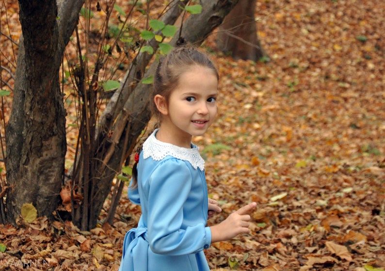 Puede incluir: Una ni&ntilde;a joven con cabello largo y casta&ntilde;o que lleva un vestido azul claro con un cuello de encaje blanco. Est&aacute; de pie en un bosque con hojas ca&iacute;das en el suelo.