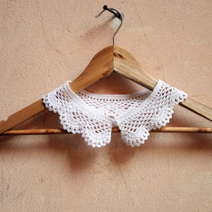 Puede incluir: Un cuello de crochet blanco con borde festoneado cuelga de una percha de madera contra una pared de color marr&oacute;n claro.