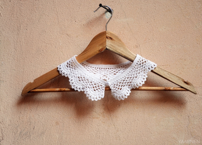 Puede incluir: Un cuello de crochet blanco con borde festoneado cuelga de una percha de madera contra una pared de color marr&oacute;n claro.
