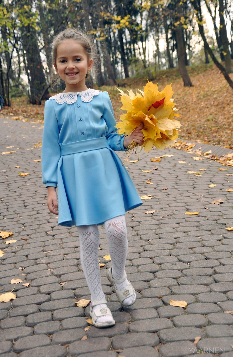 Puede incluir: Una ni&ntilde;a peque&ntilde;a con un vestido azul claro con un cuello de encaje blanco sostiene un ramo de hojas de oto&ntilde;o amarillas. Lleva medias blancas y zapatos blancos. La ni&ntilde;a est&aacute; de pie en un camino de adoquines.