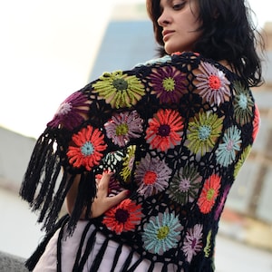 Può includere: Un top in crochet nero con motivi floreali colorati e frange. Il top è realizzato con un motivo a fiore all'uncinetto in tonalità di rosso, verde, viola e blu.