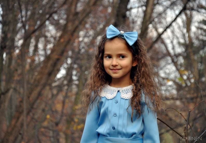 Puede incluir: Una ni&ntilde;a joven con cabello largo y casta&ntilde;o lleva un vestido azul claro con un cuello de encaje blanco y un lazo azul claro en el cabello. Est&aacute; de pie frente a un fondo borroso de &aacute;rboles.