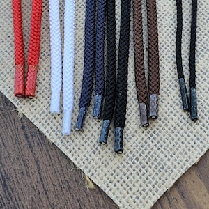 May include: A collection of ten braided shoelaces in various colors including red, white, navy blue, black, and brown. Each lace features a matching plastic aglet at the tip, providing a secure finish for these durable, round-cord style footwear accessories.