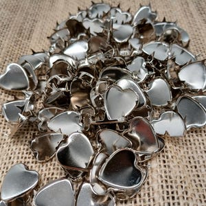 May include: A pile of shiny, silver-colored heart-shaped metal embellishments. Each heart has a small, pointed prong on the back. The hearts are scattered on a textured, tan fabric background.