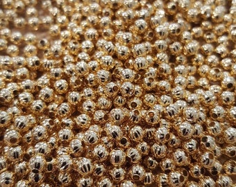 May include: A large pile of gold-colored round beads with a textured surface. The beads are scattered on a beige fabric background.