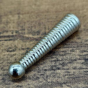 Op de afbeelding: Een zilverkleurig, cilindrisch object met een bal aan &eacute;&eacute;n uiteinde en een geribbeld, spiraalvormig ontwerp langs het lichaam. Het object ligt op een bruin, getextureerd oppervlak.