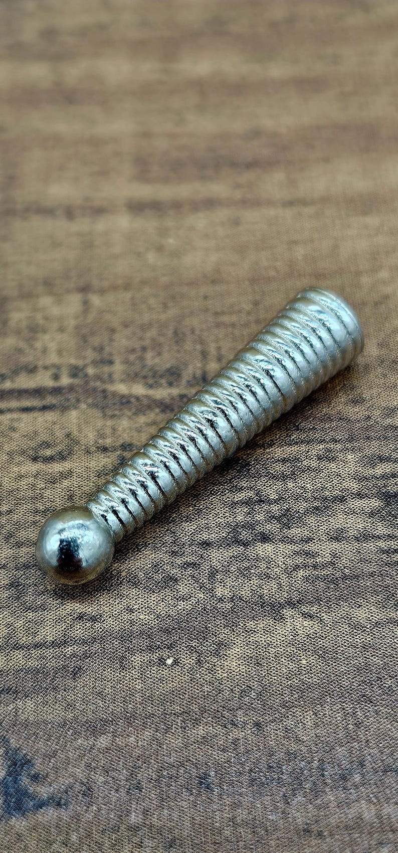 Op de afbeelding: Een zilverkleurig, cilindrisch object met een bal aan &eacute;&eacute;n uiteinde en een geribbeld, spiraalvormig ontwerp langs het lichaam. Het object ligt op een bruin, getextureerd oppervlak.
