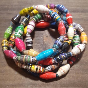 Op de afbeelding: Een kleurrijke ketting gemaakt van gerolde papier kralen. De kralen zijn in verschillende kleuren, waaronder rood, geel, blauw, groen en zwart. De kralen zijn aan elkaar geregen met zilverdraad.