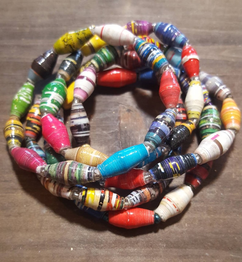 Op de afbeelding: Een kleurrijke ketting gemaakt van gerolde papier kralen. De kralen zijn in verschillende kleuren, waaronder rood, geel, blauw, groen en zwart. De kralen zijn aan elkaar geregen met zilverdraad.