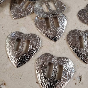 May include: A pile of silver heart-shaped metal buckles with a decorative etched pattern and a rectangular cut-out in the center.