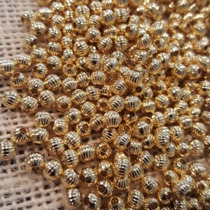May include: A large pile of gold-colored metal beads with a textured surface. The beads are round and have a small hole in the center.