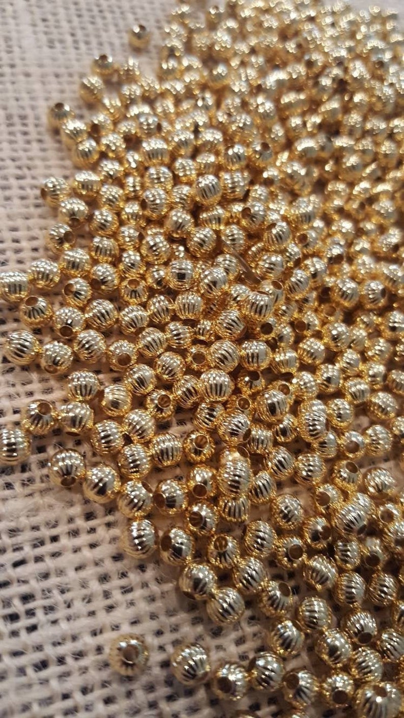 May include: A large pile of gold-colored metal beads with a textured surface. The beads are round and have a small hole in the center.
