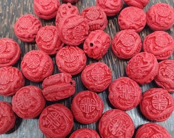 Può includere: Una collezione di piccole perline rotonde rosse con intricati disegni intagliati. Ogni perlina presenta un motivo dettagliato, forse un simbolo o un emblema, in una vibrante tonalità di rosso. Le perline sono sparse, mostrando le loro singole incisioni e trame.